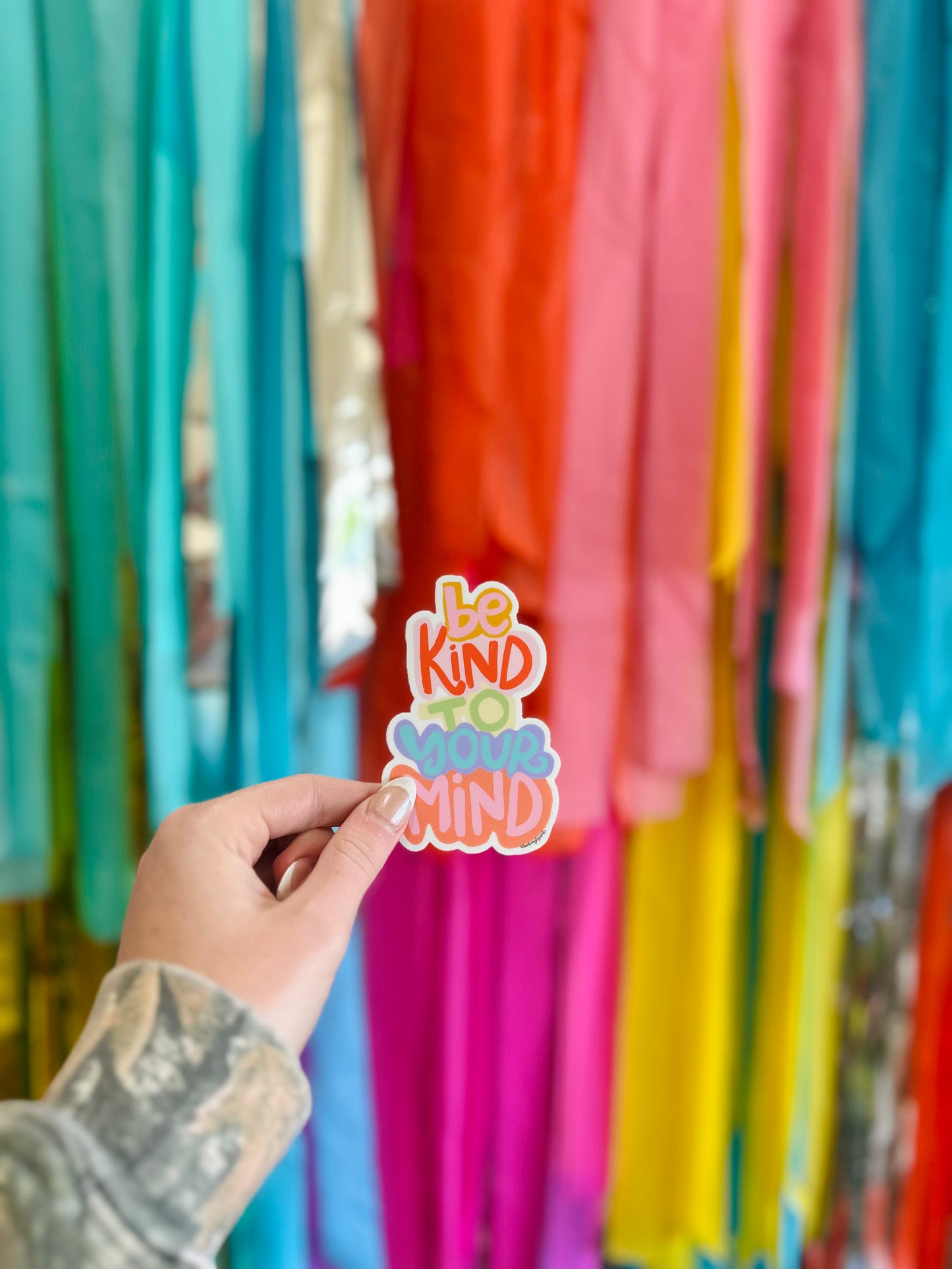 A sticker with the phrase 'Be Kind to Your Mind' written on it, held in front of a colorful backdrop with streamers.