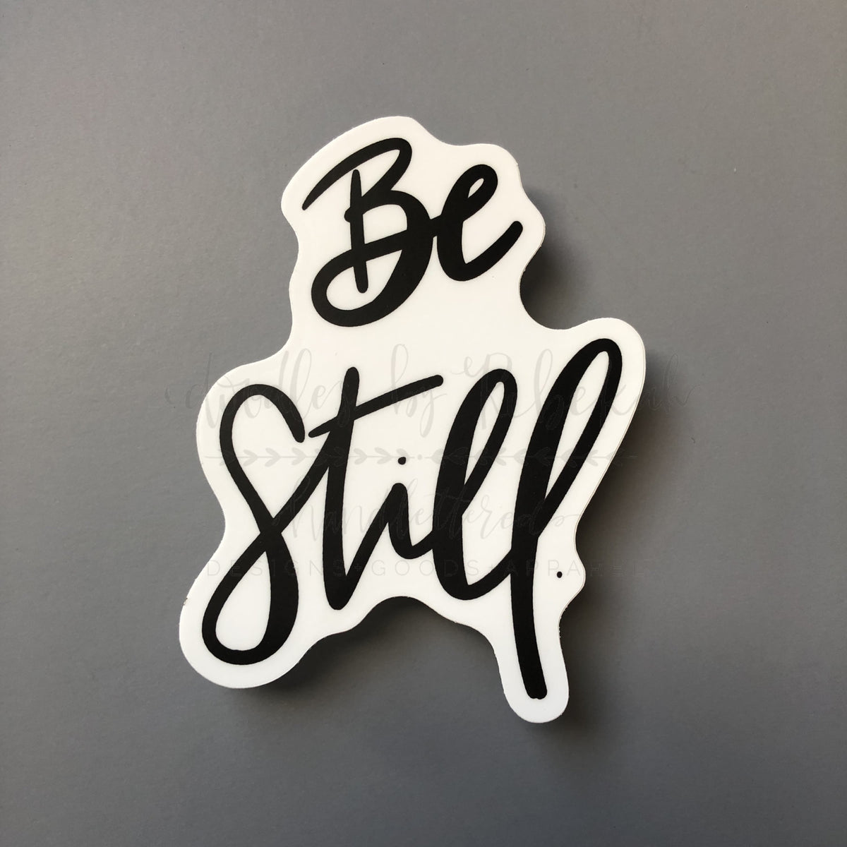 A white sticker with the phrase 'Be Still' written in a flowing black script font.