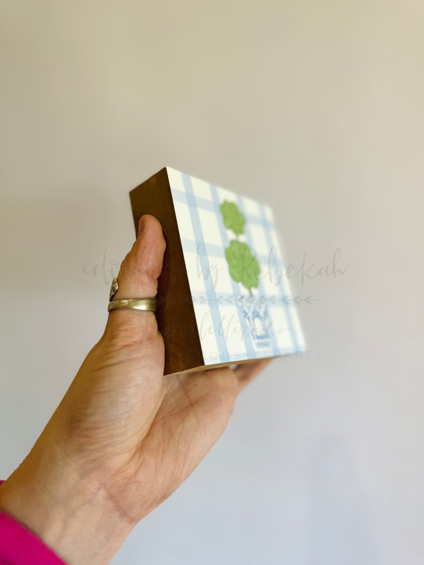 A hand holding a small decorative block with a green tree on a blue and white checkered background, with the text 'Boxwood' visible on the block.