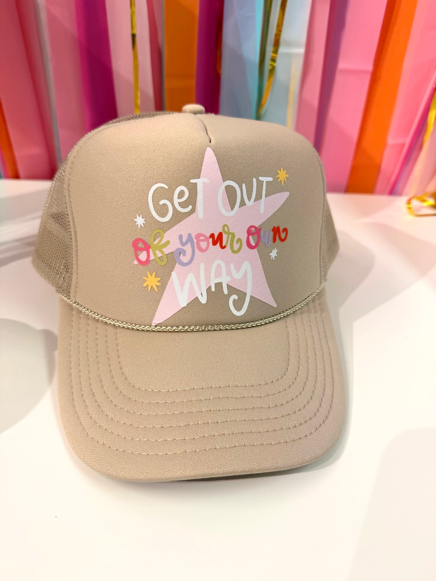 Tan "Get Out of Your Own Way" trucker hat from Doodles by Rebekah, featuring a pink star and whimsical hand-drawn typography against a colorful party streamer backdrop.