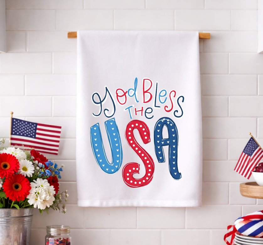 Patriotic "God Bless the USA" decorative kitchen tea towel with red, white, and blue hand-lettering and heart patterns, displayed against a white tile backsplash.