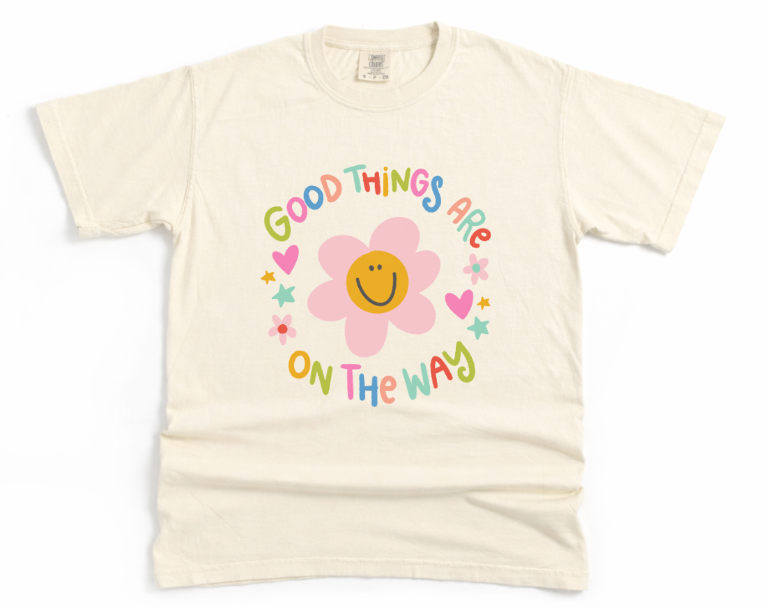 Beige t-shirt with a colorful graphic and text 'Good things are on the way' featuring a flower design.