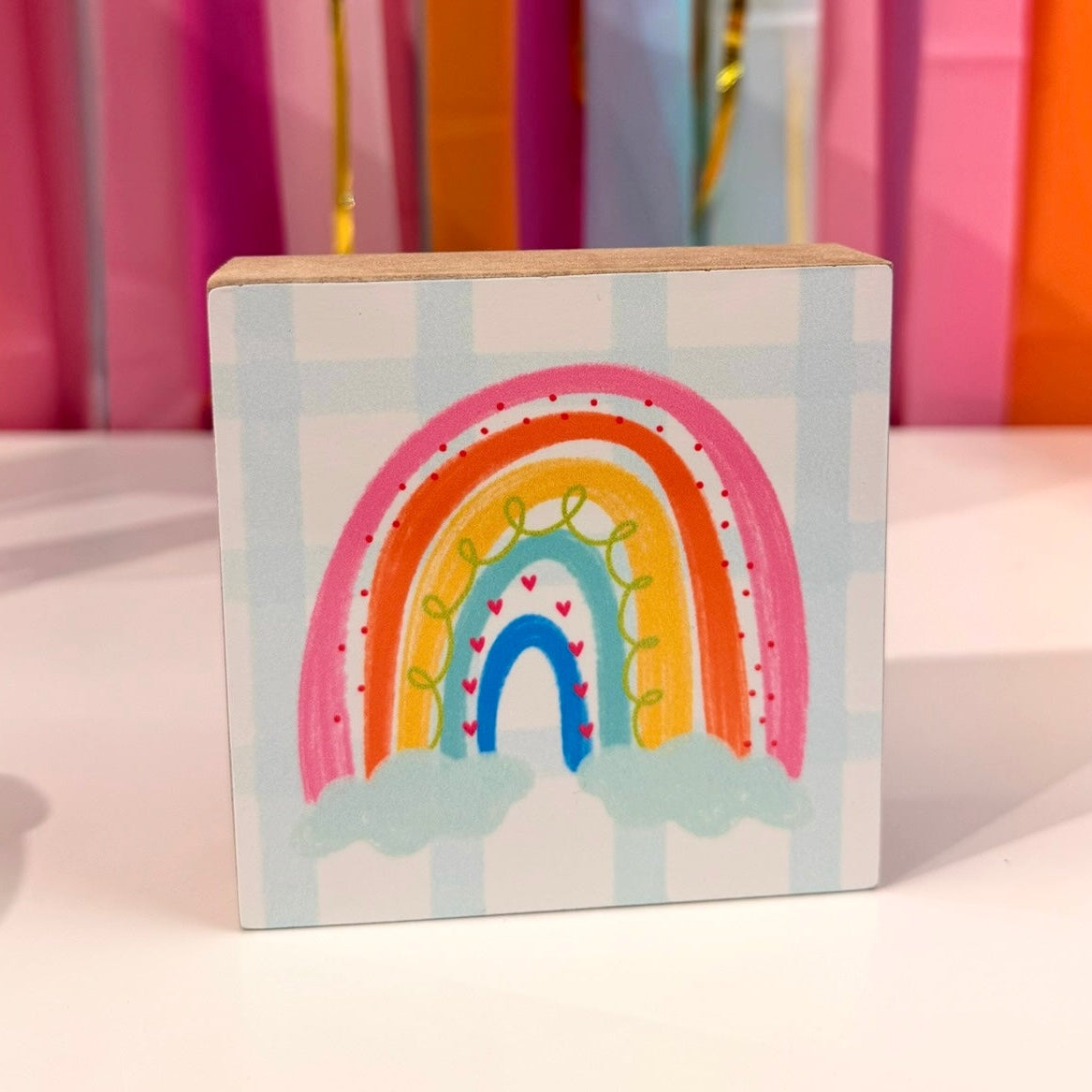 Doodles by Rebekah rainbow wooden block home decor featuring a whimsical hand-painted colorful rainbow and checkered pattern. This cheerful tabletop or nursery shelf accent is shown against a bright party streamer background.