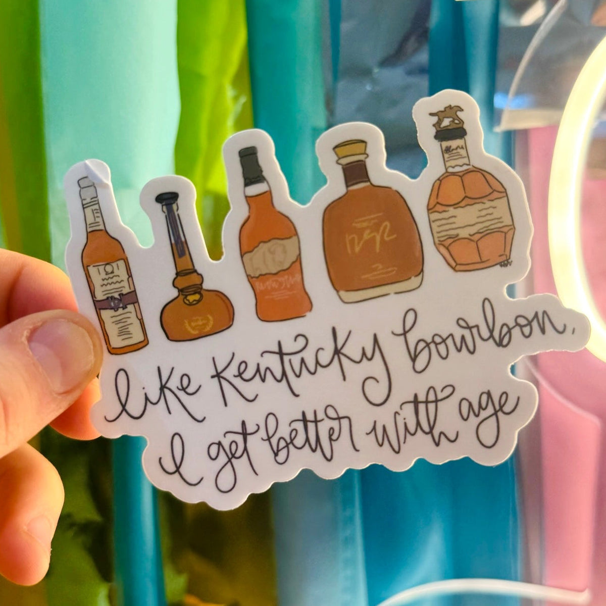 Doodles by Rebekah "Kentucky Bourbon" birthday sticker. This whimsical vinyl decal features five different bourbon bottles and an inspirational aging quote, making it a perfect birthday gift for bourbon lovers to display on water bottles, laptops, and home bars.