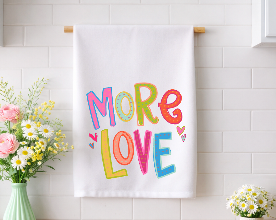 Whimsical "MORE LOVE" decorative tea towel with vibrant, hand-lettered typography and heart patterns, displayed in a bright kitchen with fresh floral arrangements.