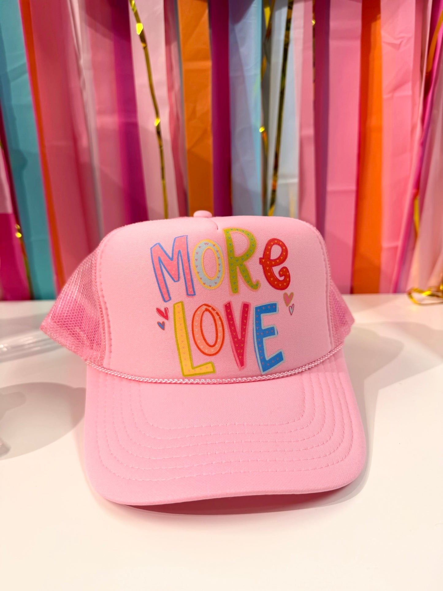 Pink "More Love" trucker hat from Doodles by Rebekah, featuring whimsical hand-drawn typography and heart illustrations on a colorful party streamer background.