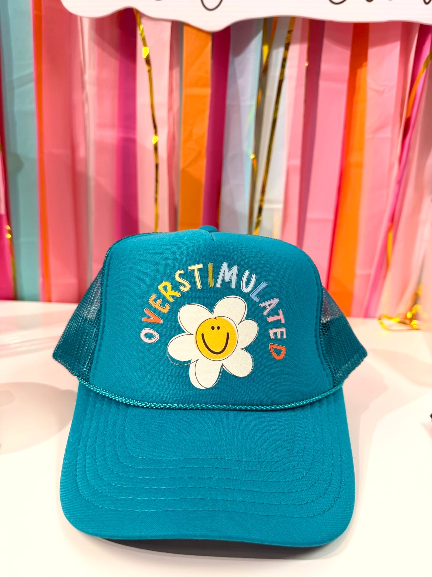 Doodles by Rebekah "Overstimulated" teal trucker hat featuring a whimsical hand-drawn daisy smiley face and colorful arched typography on a party streamer background.
