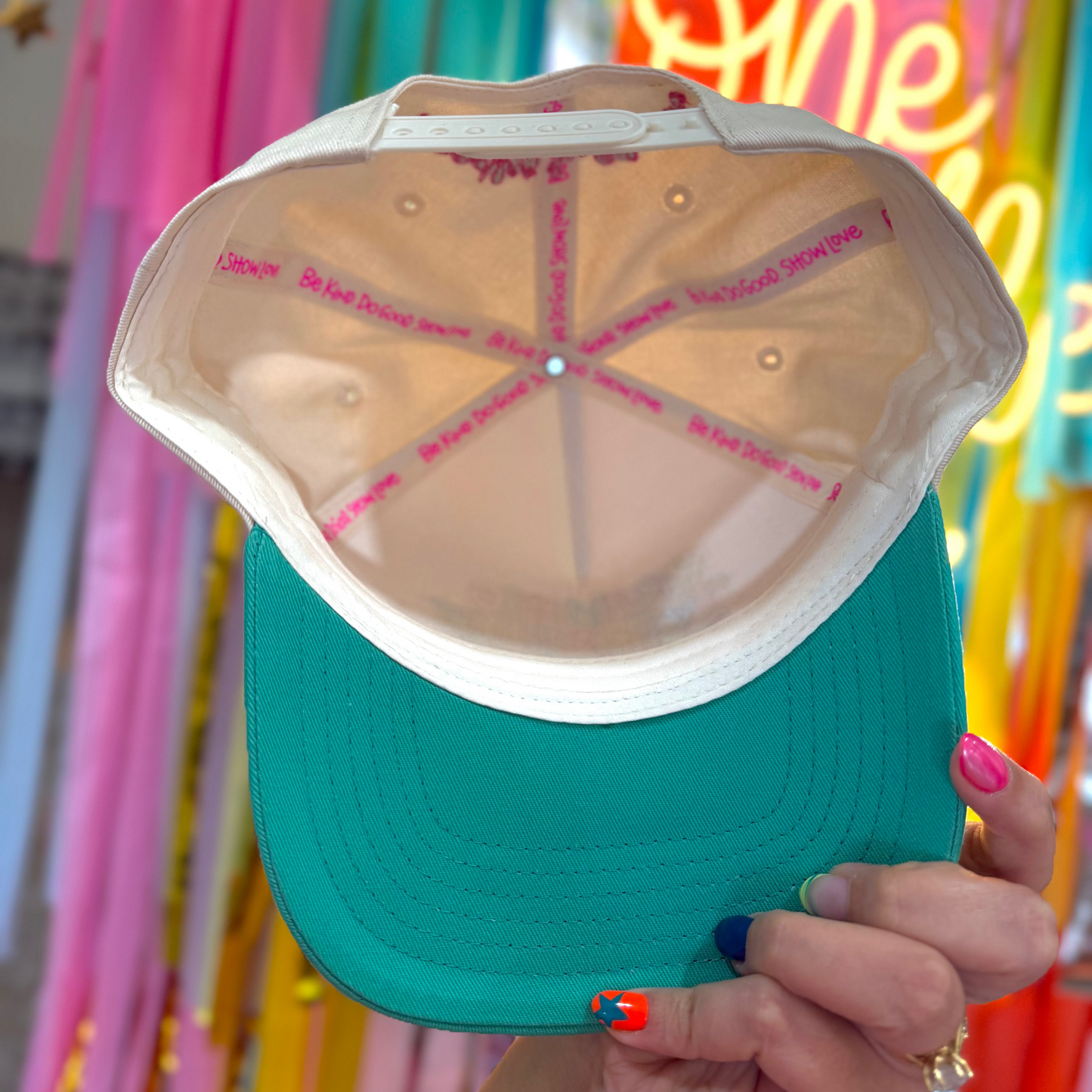 Teal cap with pink text held in front of colorful balloons and decorations
