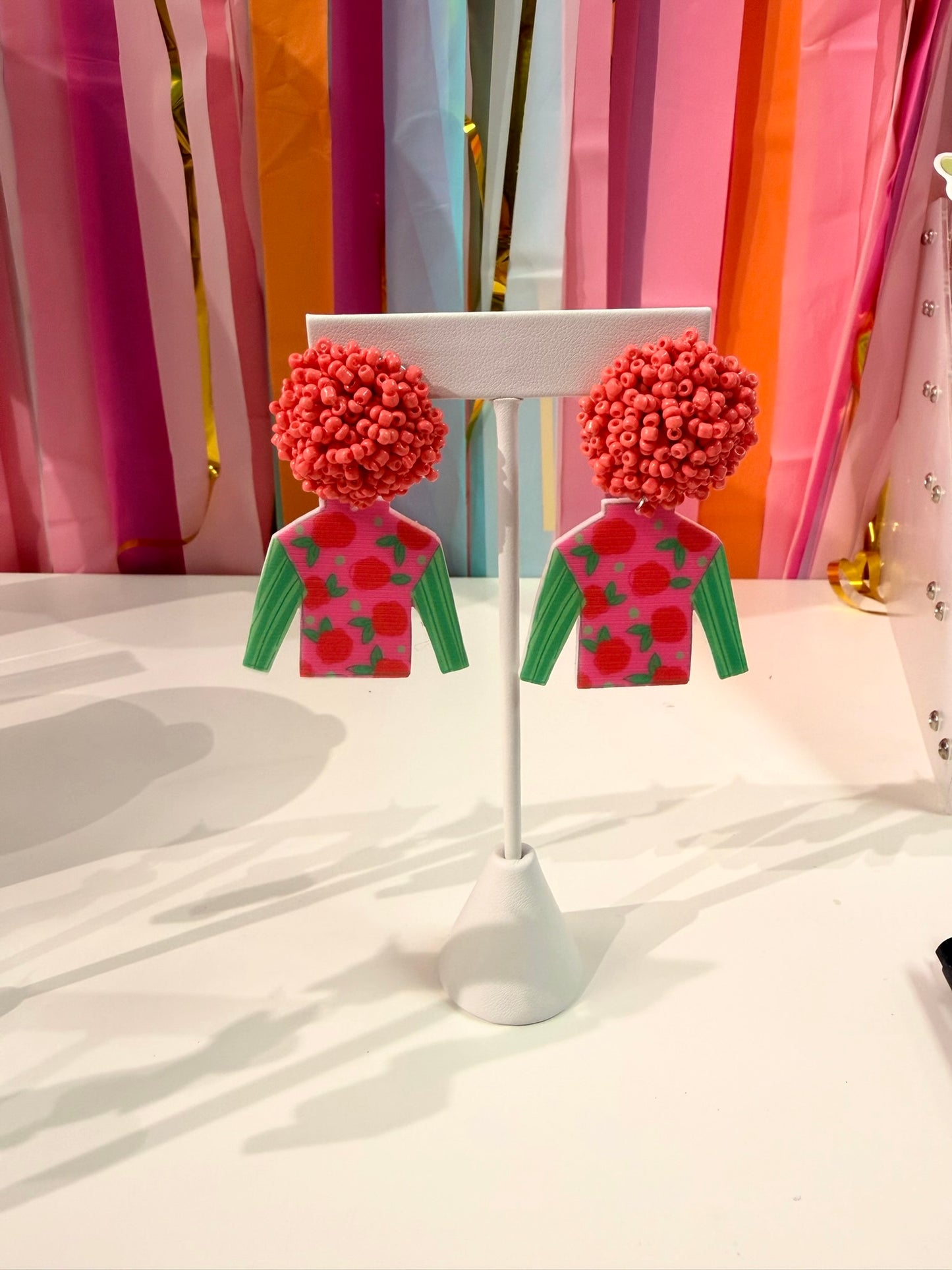 Doodles by Rebekah Kentucky Derby jockey silk earrings featuring hand-drawn rose patterns, green sleeves, and pink beaded studs. These whimsical horse racing accessories are shown against a bright party streamer backdrop.