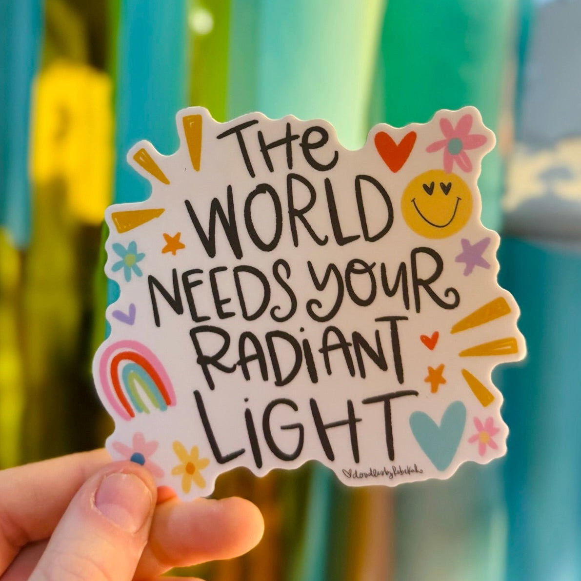 Doodles by Rebekah "The World Needs Your Radiant Light" inspirational vinyl sticker. This whimsical hand-painted motivational decal with rainbow and smiley face accents is a perfect gift for decorating water bottles, laptops, and mirrors to spread positivity.