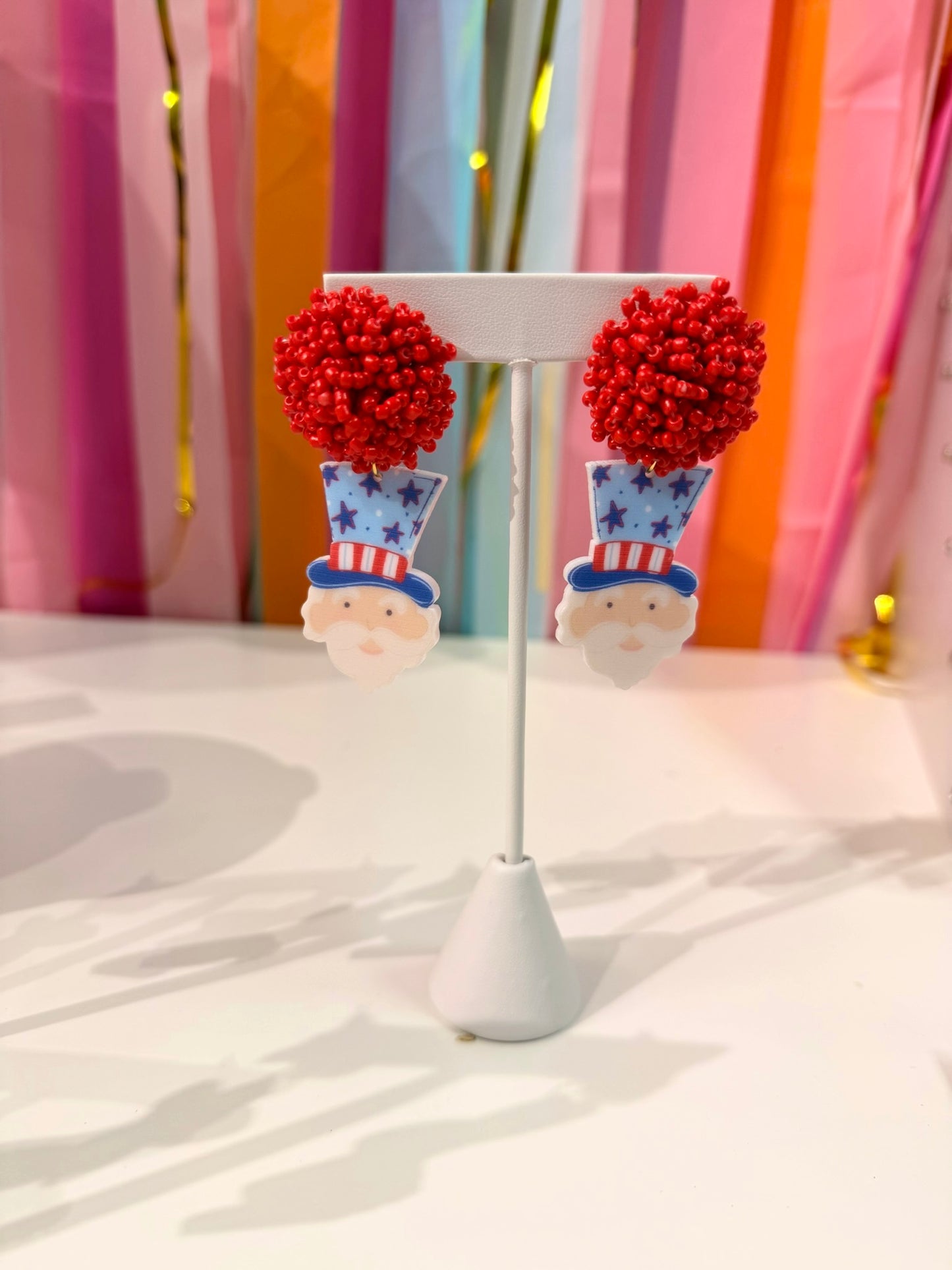 Doodles by Rebekah Uncle Sam statement earrings featuring hand-painted patriotic designs and red beaded studs. These whimsical Fourth of July accessories are shown against a bright party streamer backdrop.