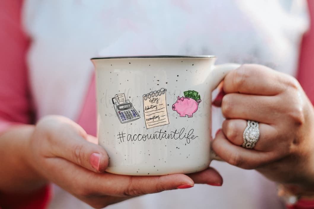 A hand holding a white ceramic mug with an illustration of accounting-themed items like a calculator, paperwork, and a pink fruit, along with the text '#ACCOUNTANTLIFE'.