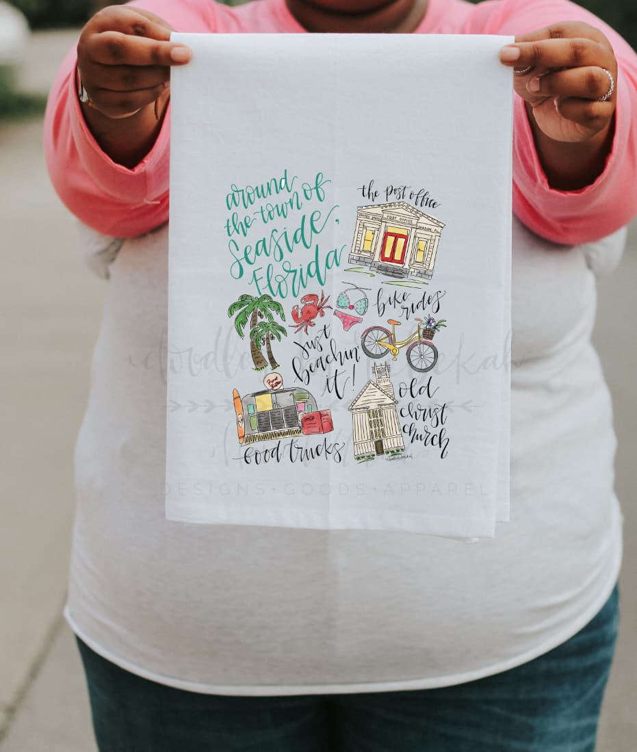 A person holding a white tea towel with a colorful illustrated design representing 'Around Seaside Florida'.