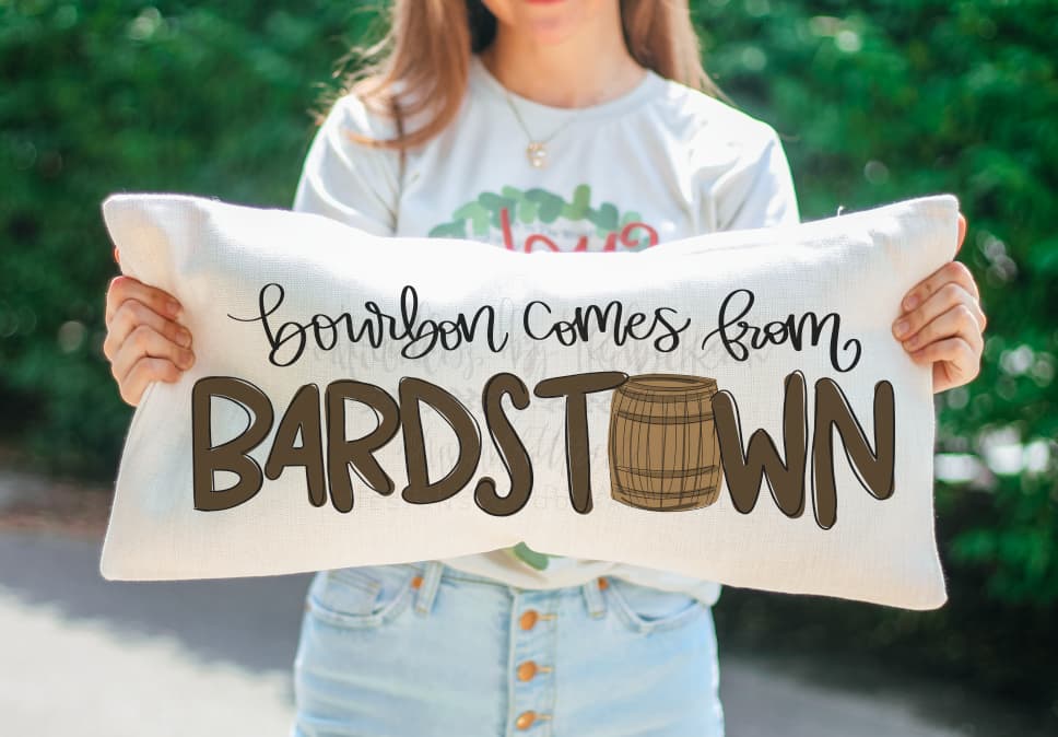 A person holding a beige lumbar pillow with the text 'Bourbon comes from BARDSTOWN' printed on it.