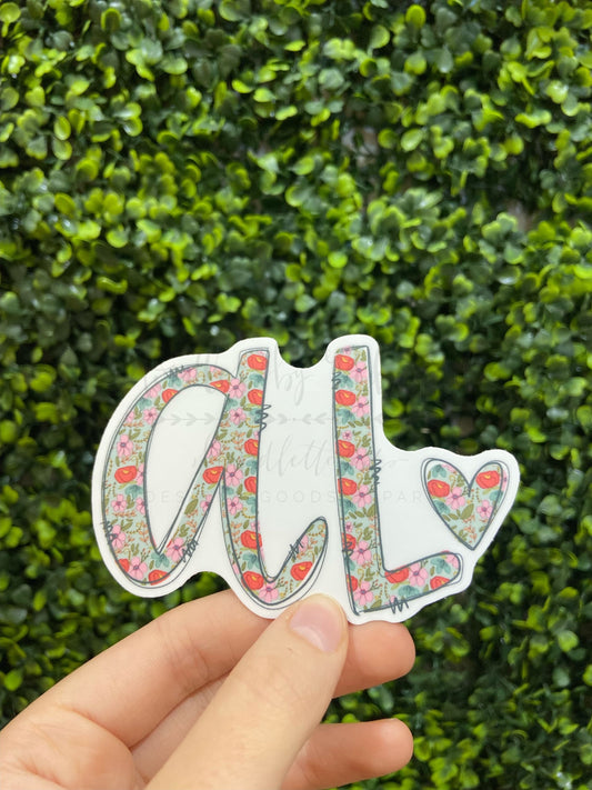A sticker with a floral pattern and a heart shape, featuring the letter 'AL' in the center, held between fingers.