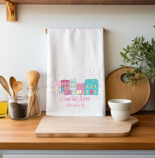A white polyester tea towel with a custom town name and logo printed in green, displayed over a kitchen counter.
