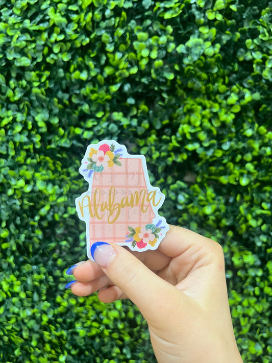 a hand holding up a pink plaid sticker in the shape of Alabama with the word Alabama in gold on it