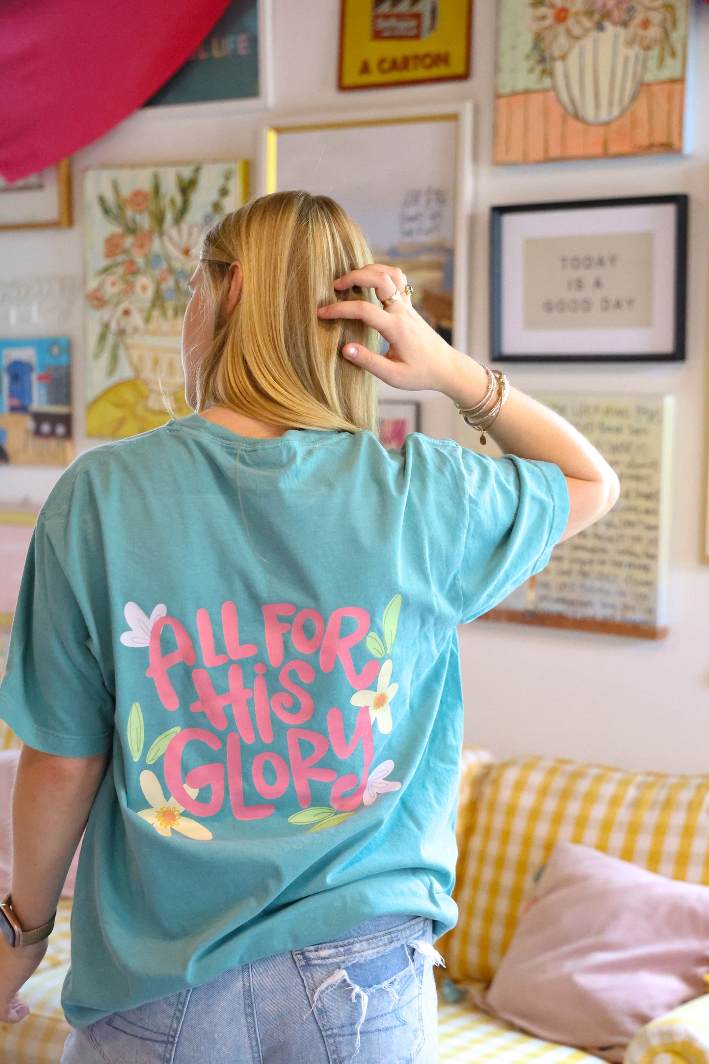 Person wearing a teal t-shirt with 'All for This Glory' text in a room with colorful decor.