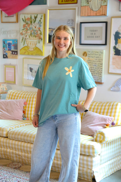 Woman wearing a blue t-shirt with a yellow flower design in a room with a checkered sofa and framed pictures on the wall.