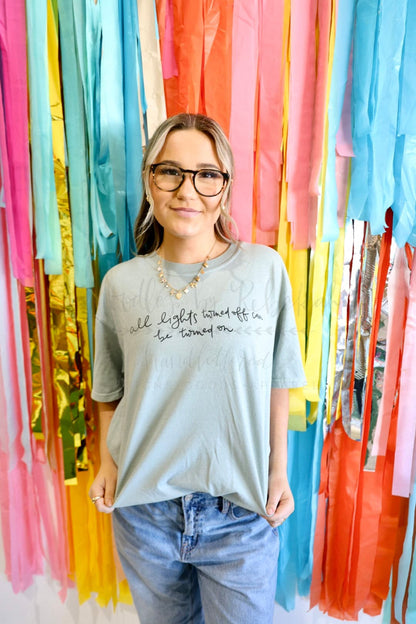 A person standing in front of a colorful backdrop, wearing a grey t-shirt with hand-drawn lettering, jeans, and glasses.