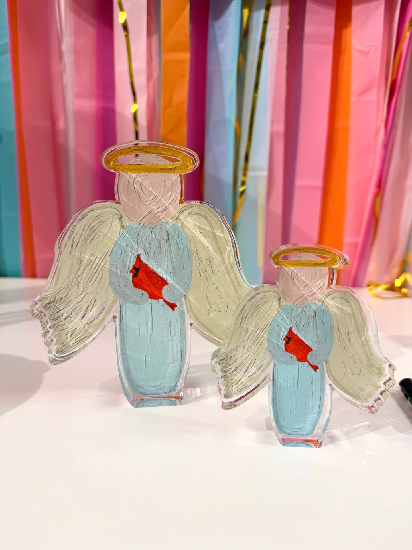 Doodles by Rebekah angel with cardinal acrylic home decor featuring whimsical hand-painted religious imagery and a red bird accent. These peaceful tabletop decorations are shown against a bright party streamer background.