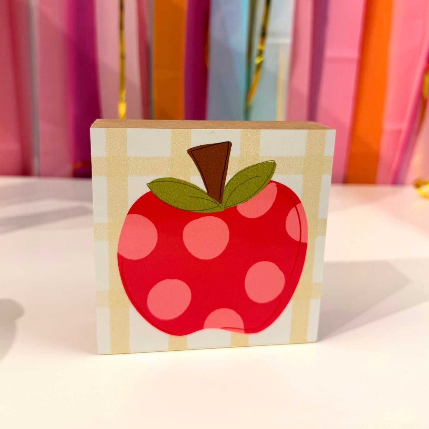 Doodles by Rebekah apple wooden block home decor featuring a whimsical hand-painted red polka dot apple on a checkered background. This charming teacher gift or kitchen tabletop accent is shown against a bright party streamer backdrop.