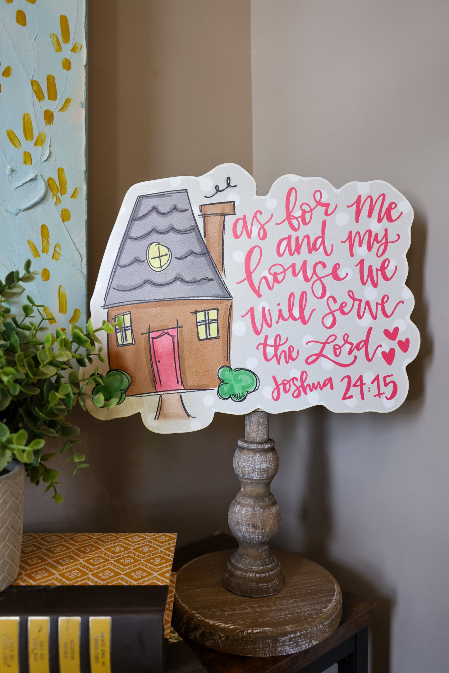 Decorative sign with house illustration and Bible verse on a stand against a wall.