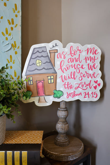Decorative sign with house illustration and Bible verse on a stand against a wall.