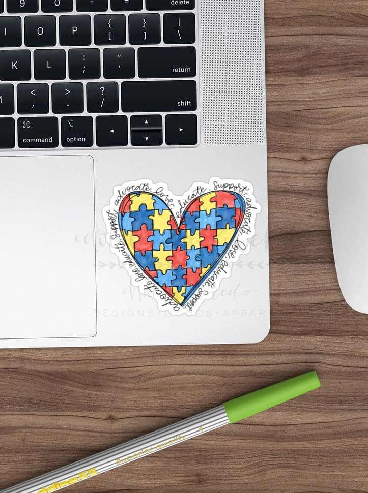 A colorful heart-shaped sticker with a autism awareness pattern placed on a laptop next to a computer mouse and a pen.