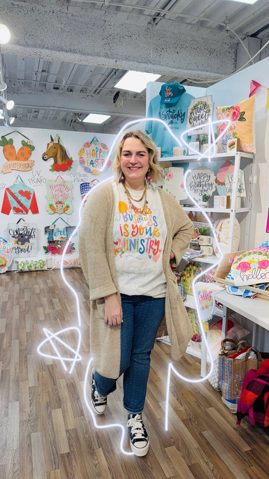 A person standing in a retail environment wearing a beige sweatshirt with the text 'YOUR BUSINESS IS YOUR MINISTRY' on it, paired with blue jeans and white sneakers.