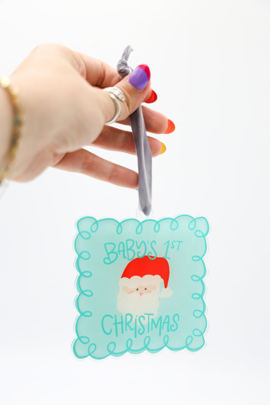 Hand holding a small ornament with a Santa Claus design and 'Baby's 1st Christmas' text on a white background.