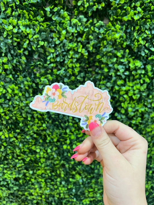 A held sticker with a floral pattern and the word 'BARDSTOWN' written on it, against a green hedge background.