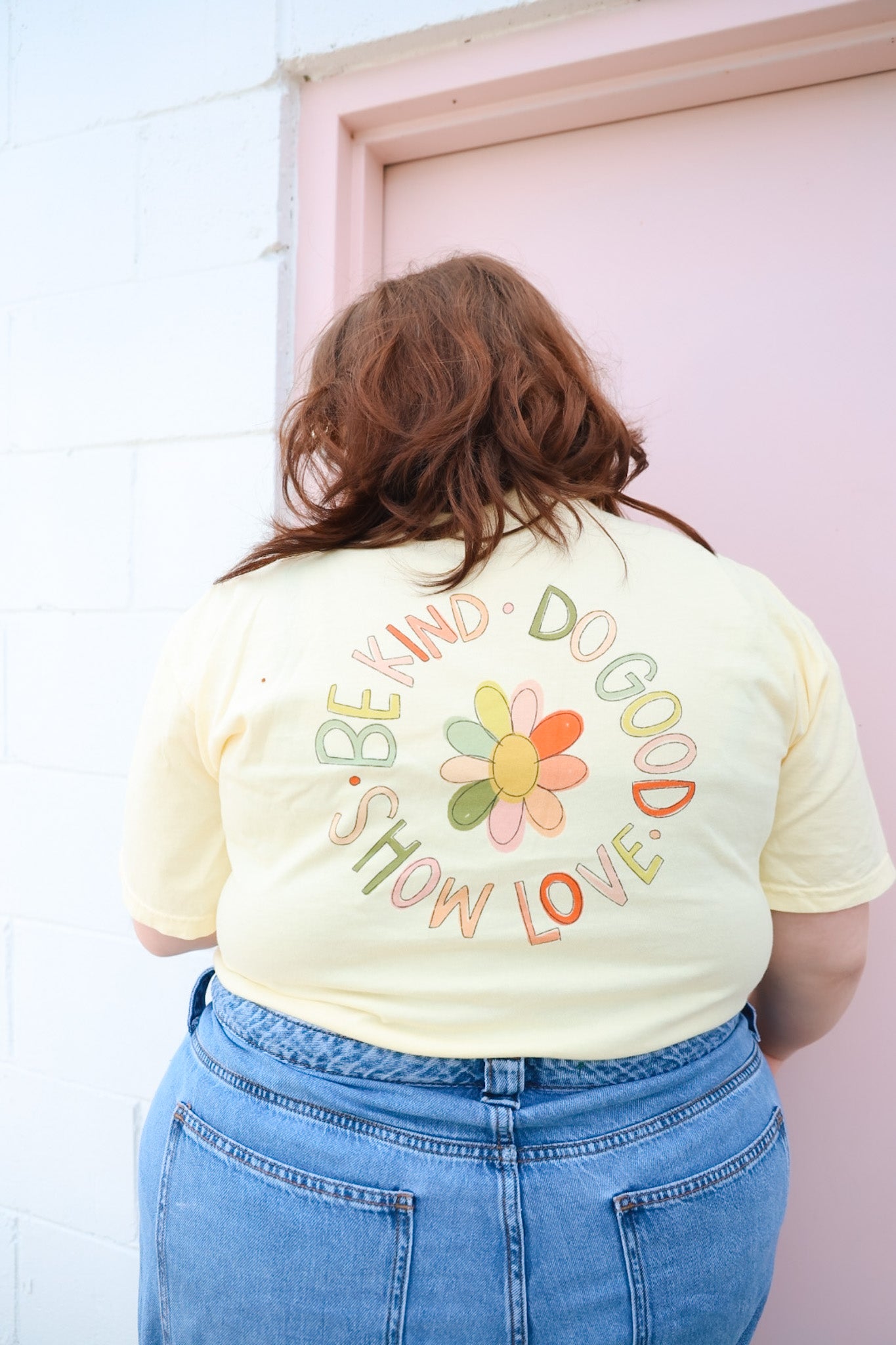 A person wearing a yellow t-shirt with hand-drawn lettering and floral designs on the back, standing in front of a pink door.