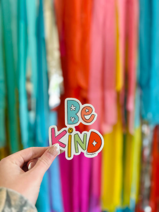 A hand holding a colorful sticker with the text 'BE KIND' written on it, with a background of colorful streamers.
