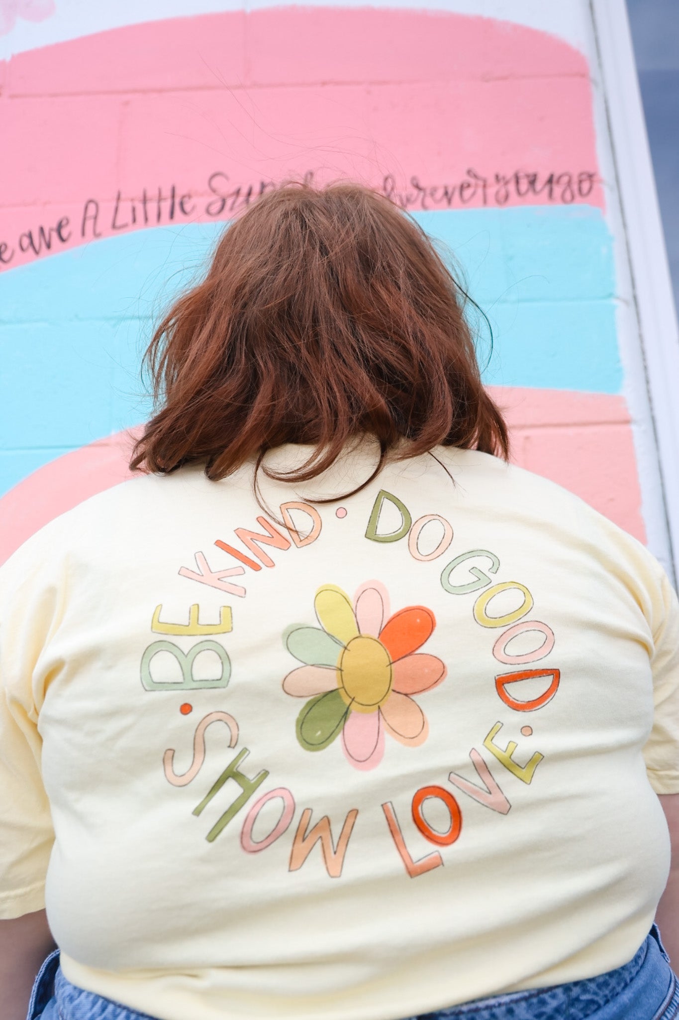 Person wearing a yellow t-shirt with graphic of flower inside a phrase reading "Be kind, do good, show love"