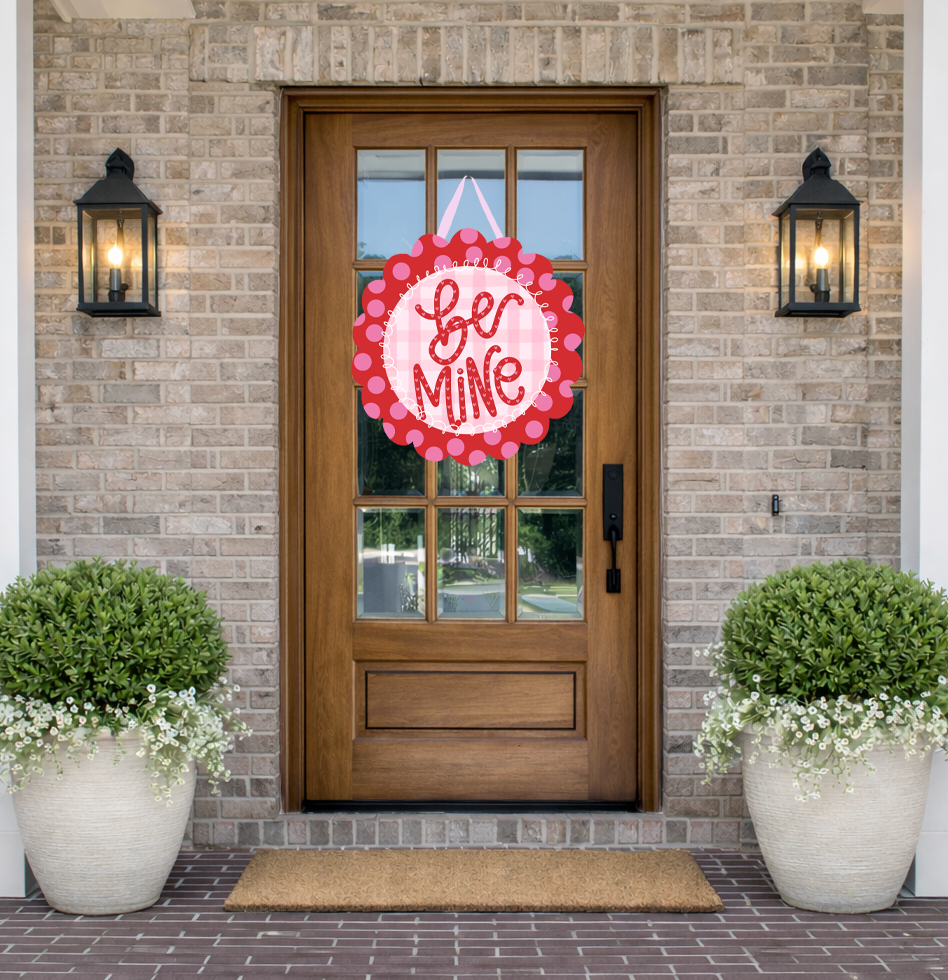 Front door with 'Be Mine' sign on a brick wall, flanked by potted plants.