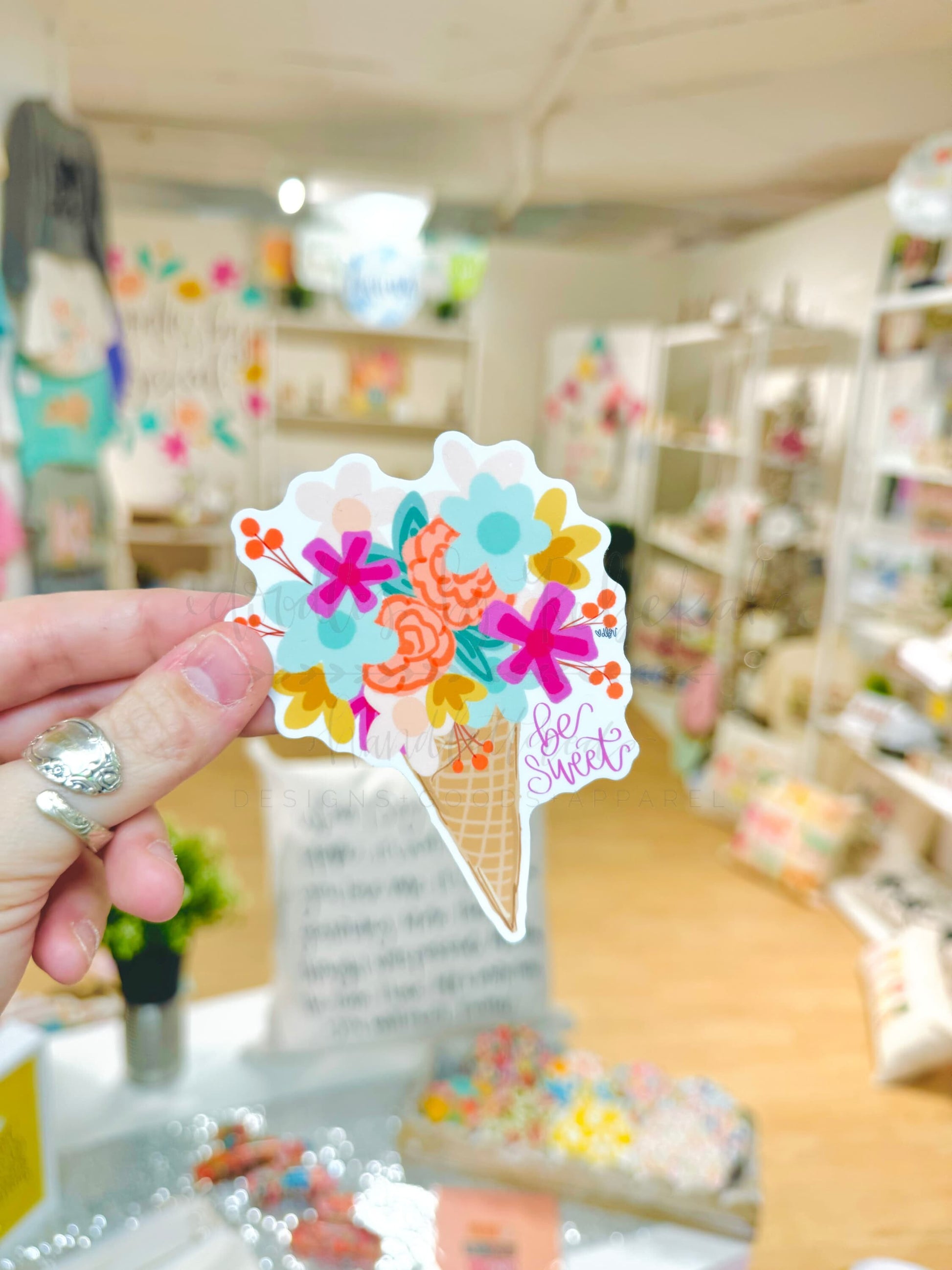 A sticker with a colorful floral design on a cone shape, held up against a blurred shop display background.
