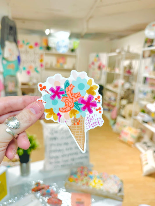 A sticker with a colorful floral design on a cone shape, held up against a blurred shop display background.