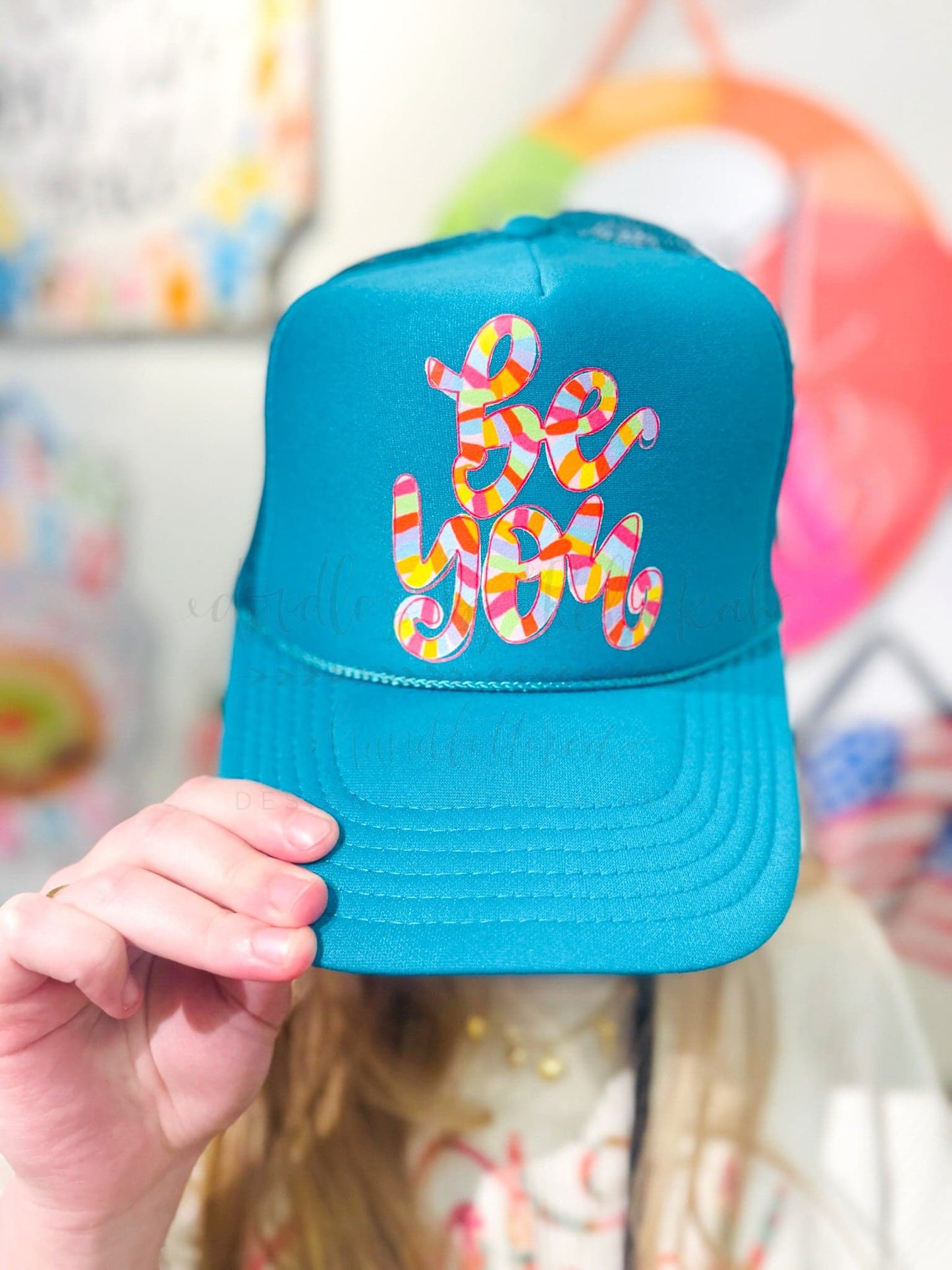 Woman wearing a teal blue trucker hat with colorful graphic 'be kind'