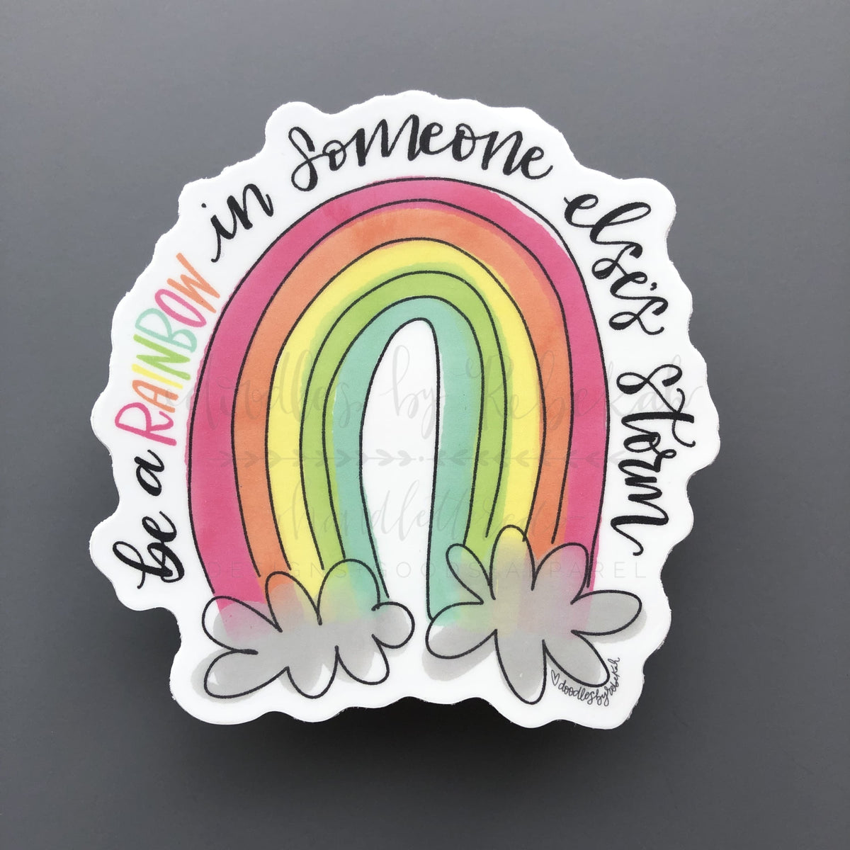 A sticker with a colorful rainbow design and text 'be a rainbow in someone else's storm' surrounded by flowers at the bottom.
