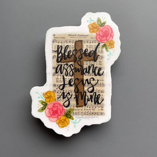 A sticker with a religious quote 'Blessed Assurance Jesus Is Mine' with a cross and floral design on a white background.