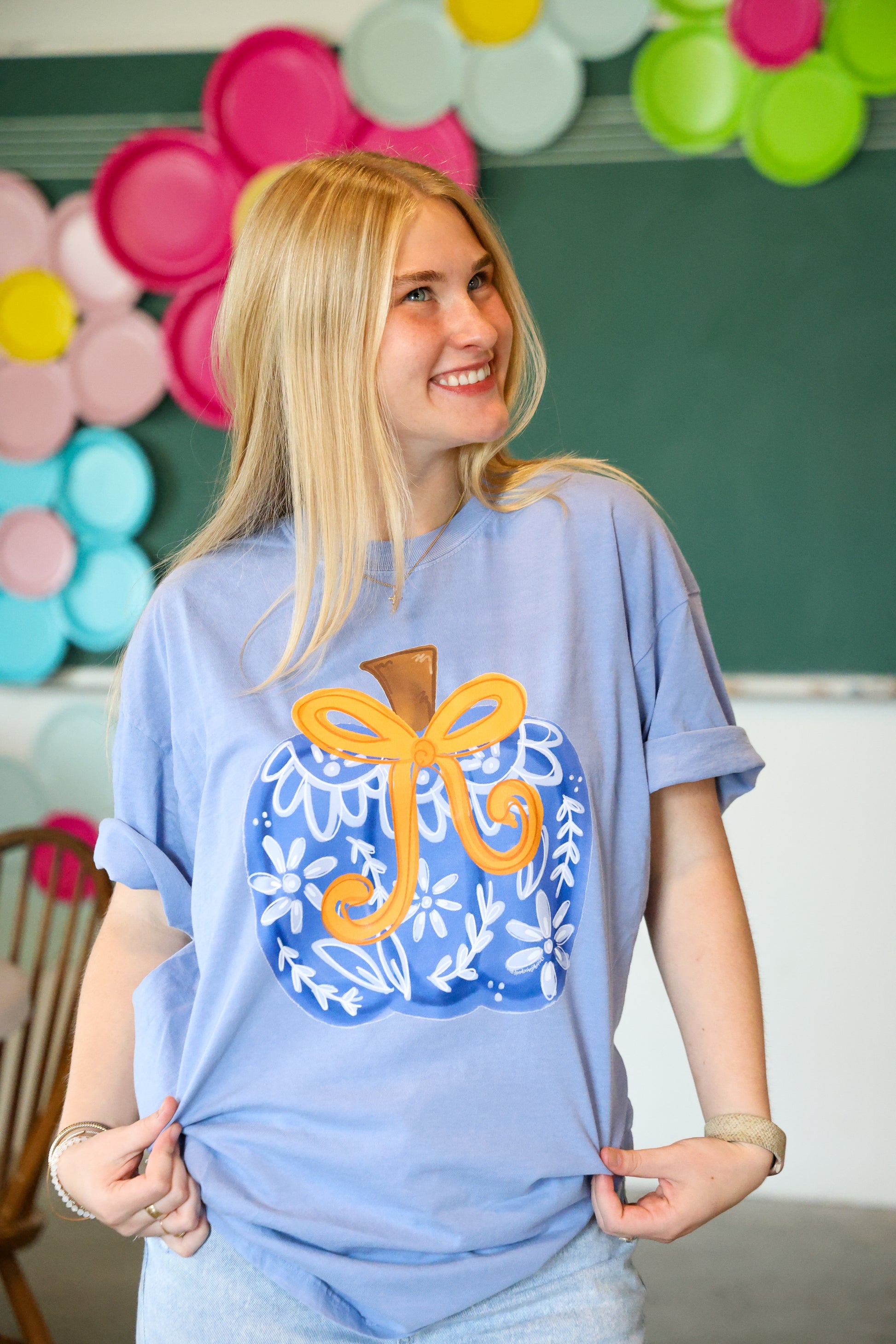 Person wearing a blue t-shirt with a decorative design, standing in front of colorful balloons and a chalkboard.