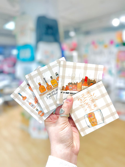 A set of four white coasters with a bourbon theme, featuring doodle-like illustrations, held in a hand.