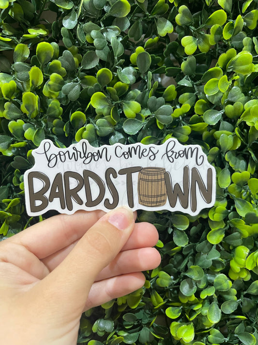A hand holding a sticker with the text 'Bourbon Comes from BARDSTOWN' placed against a leafy green background.