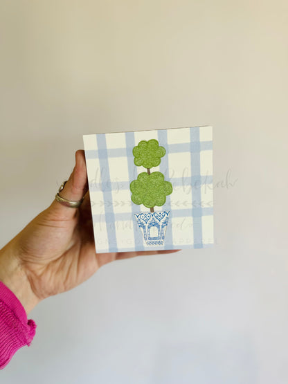 A hand holding a small decorative block with a green tree on a blue and white checkered background, with the text 'Boxwood' visible on the block.