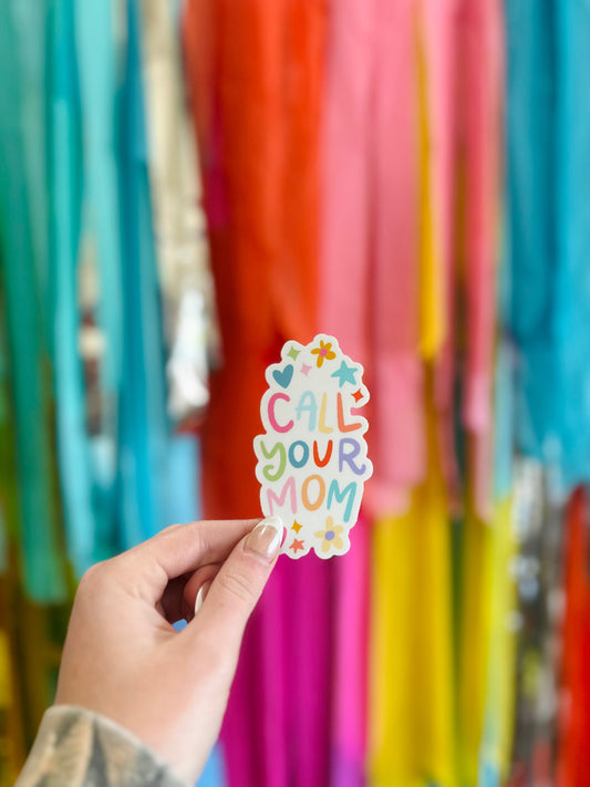 A hand holding a sticker with the text 'CALL YOUR MOM' on a colorful background with streamers.