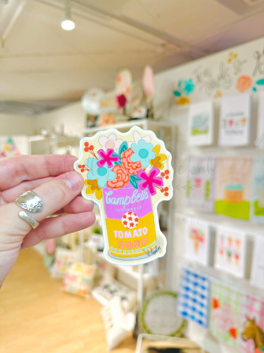A hand holding a colorful sticker with floral patterns and the text 'Campbell's Tomato Soup' on it, with a blurred background of a store.