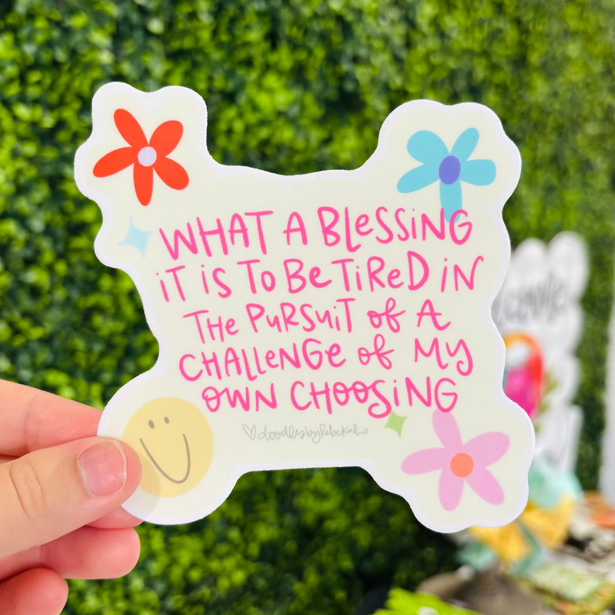 A hand holding a sticker that reads "What a blessing it is to be tired in the pursuit of a challenge of my own choosing"