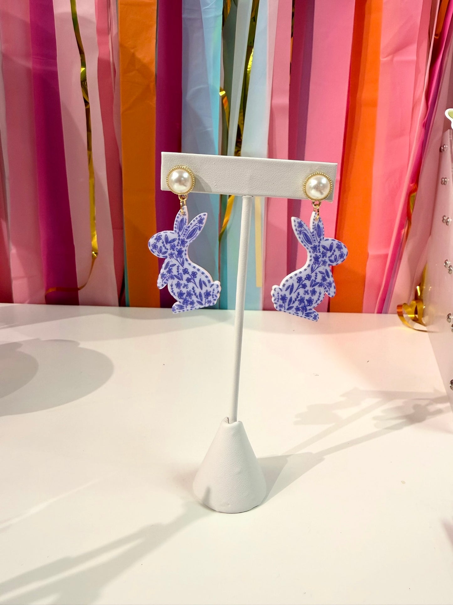 Doodles by Rebekah chinoiserie bunny earrings featuring hand-drawn navy and white floral patterns and pearl studs. These whimsical ginger jar-inspired spring accessories are shown against a bright party streamer backdrop.
