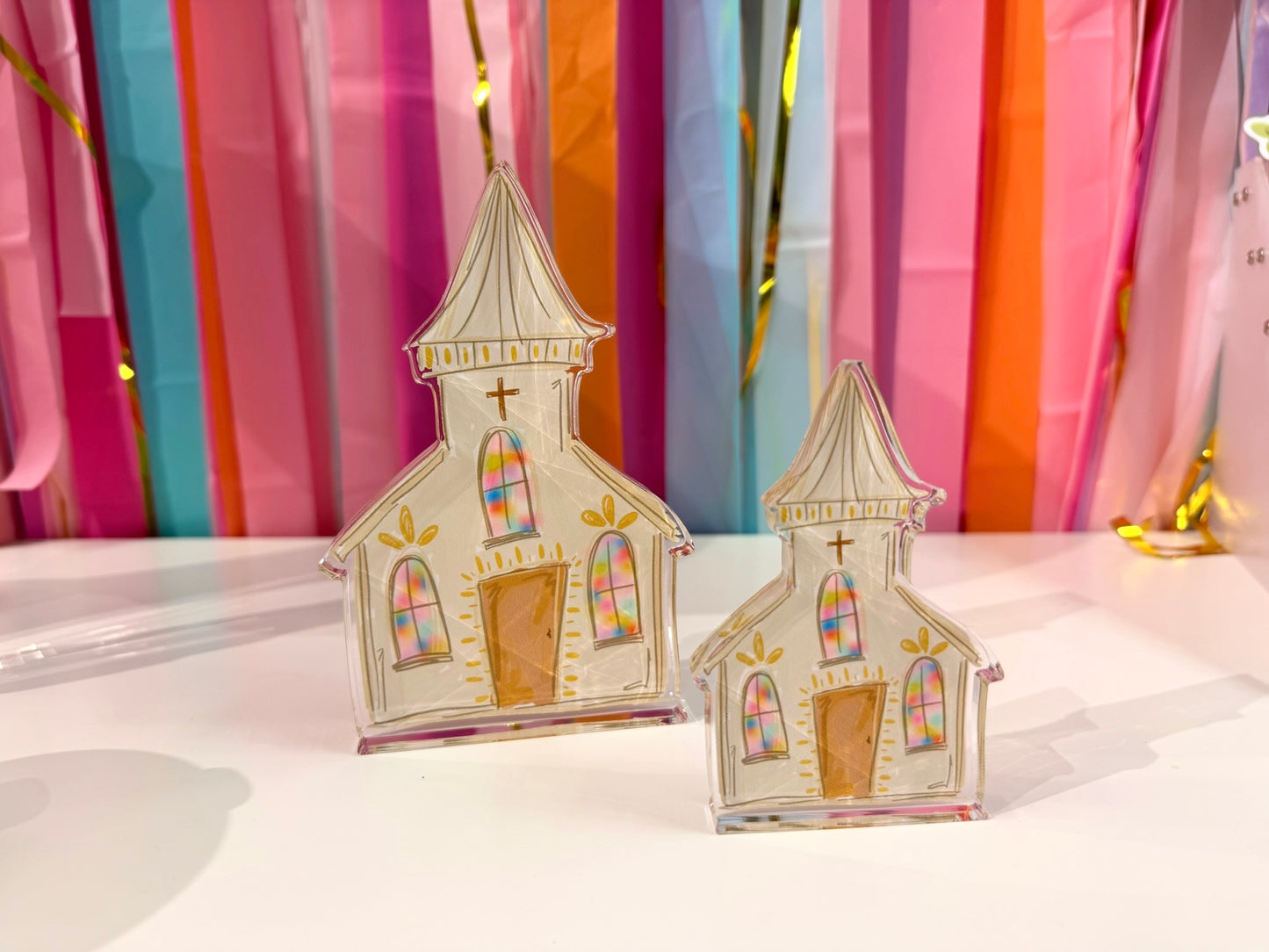 Doodles by Rebekah white church acrylic home decor featuring whimsical hand-painted stained glass windows and gold accents. These colorful religious or spring tabletop decorations are shown against a bright party streamer background.